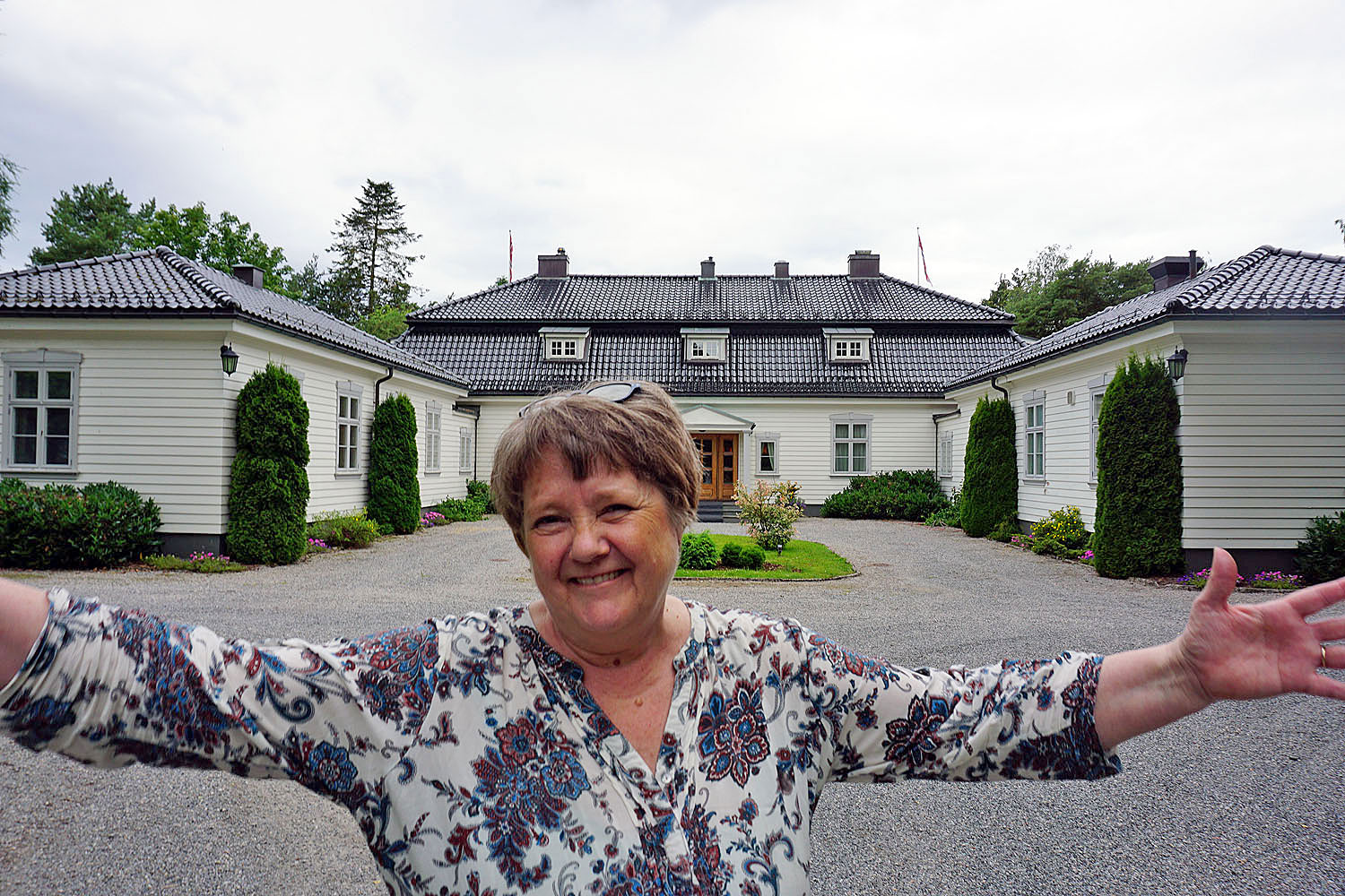 woman, arms out, welcoming, smiling, posing, white building, private but for business, garden and impressive main entrance.