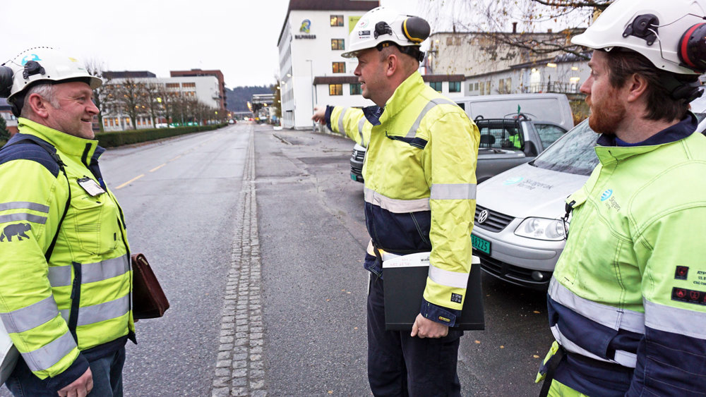 tre personer står i gate, en person peker nedover gaten. Alle har gule jakker og hvite hjelmer. I industripark.