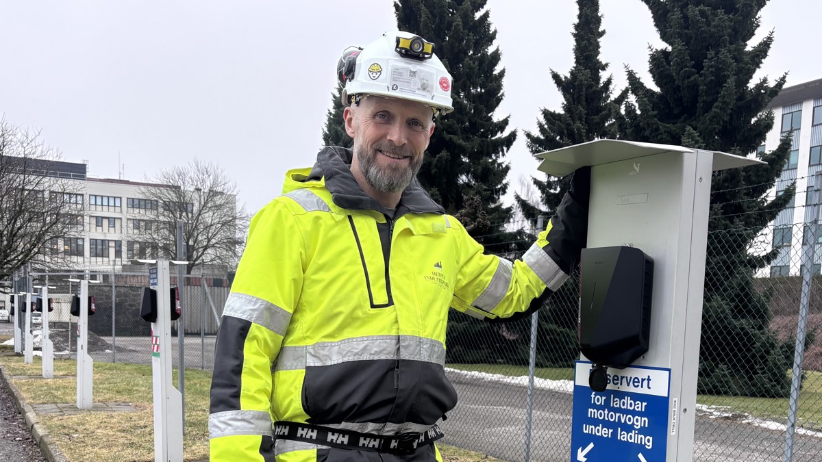 mann, poserer, står ved elbil ladestasjon, hvit hjelm, gul jakke, fabrikkgjerde