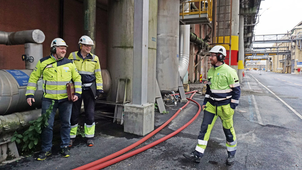 tre personer utenfor bygg i prosessindustri, rørledninger på bakken.