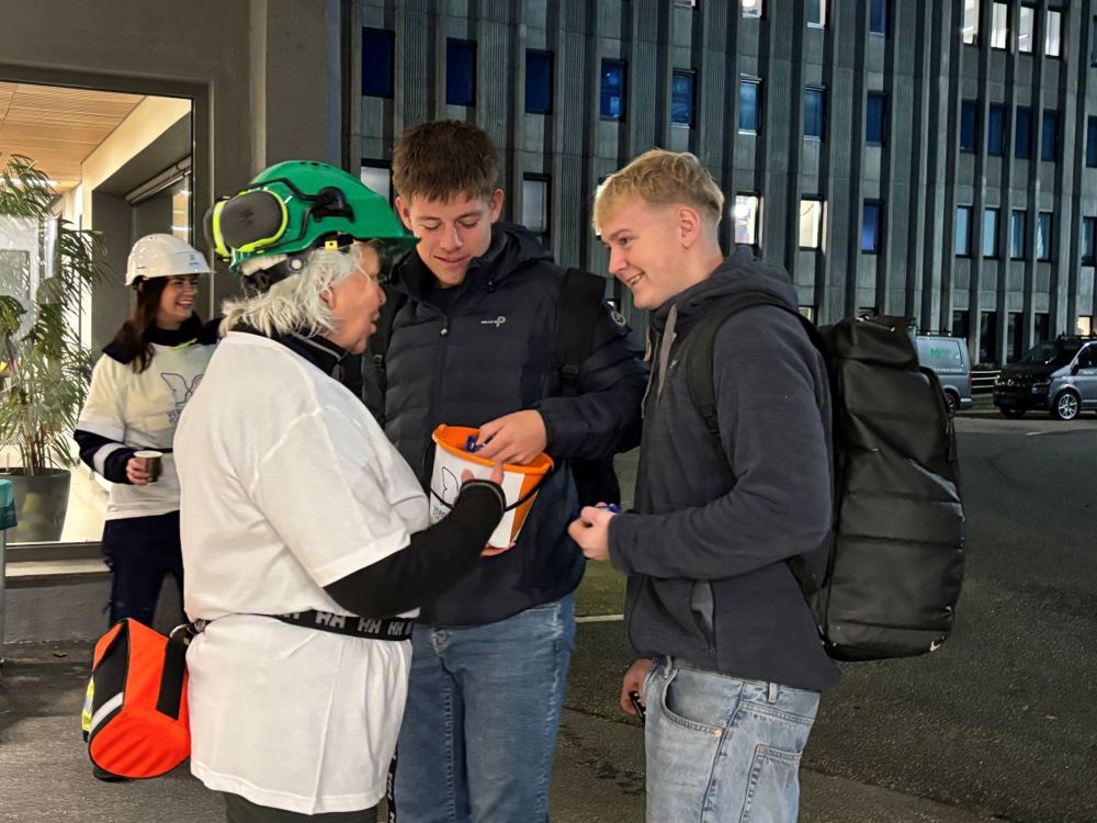 to unge menn plukker karameller fra en oransje bøtte, person med hvite tskjorte og grønn hjelm deler ut.