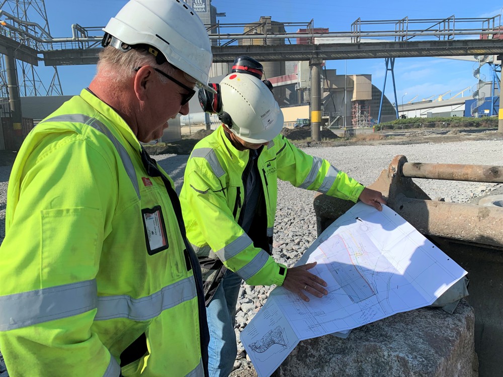 to personer står ute på et stort anleggsområde og studerer en stor plantegning. Området har såle av pukk.