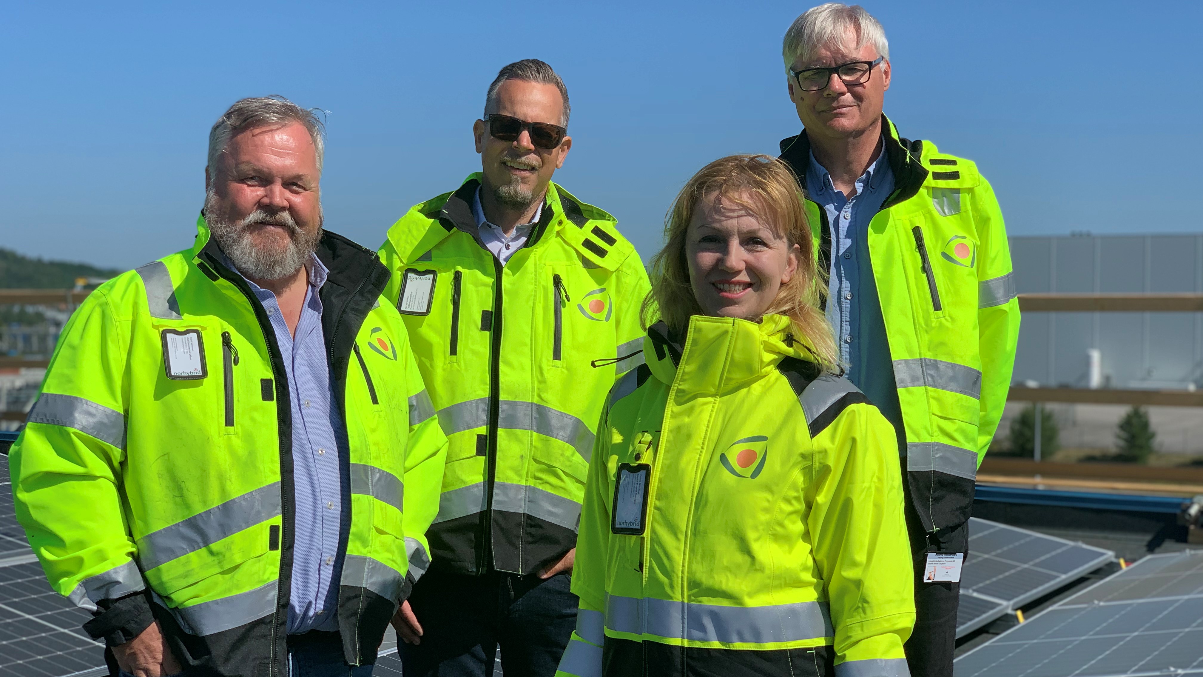 four persons, three men and one woman standing on a flat rooftop. They are wearing yellow jackets. sunpanels on the roof.