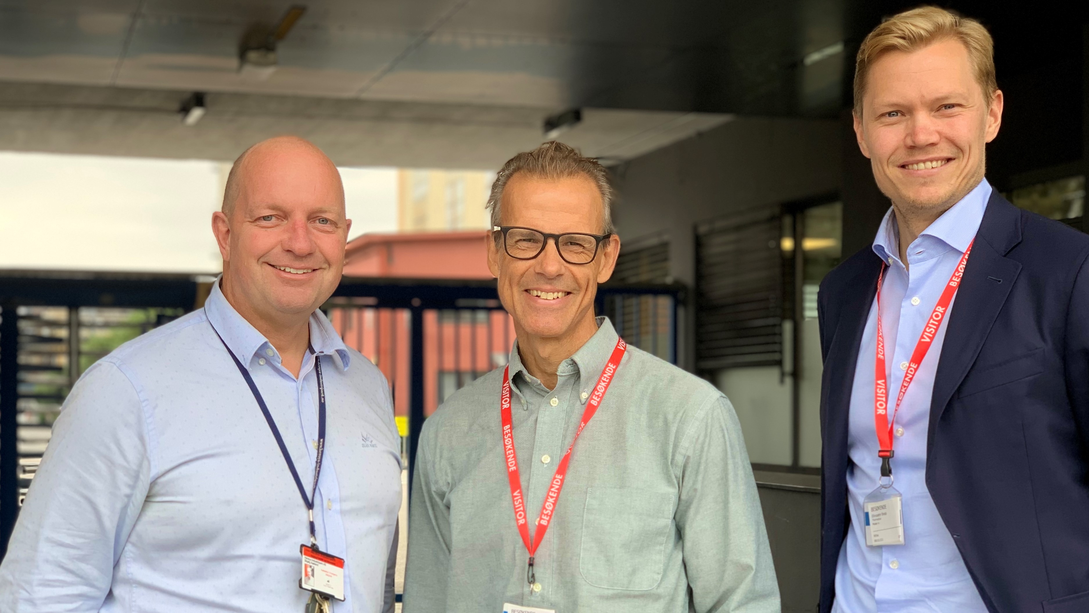 three men standing next to eachother, posing, wearing different shades of light blue shirts, key-card around their necks, standing near entrance gate to industrial park.