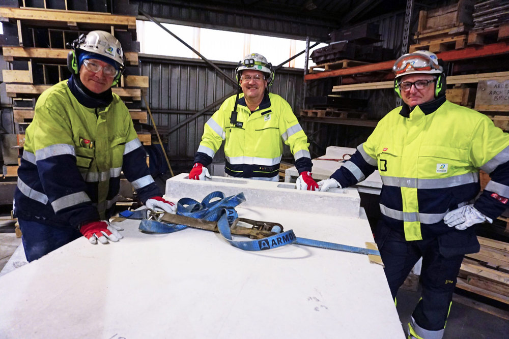 tre menn poserer, står ved arbeidsbord med ildfaste kasser, arbeidstøy, verneutstyr