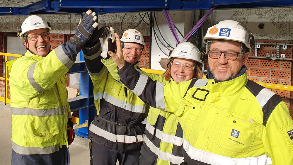 four people high-five, yellow jackets, white helmets, production plant area.