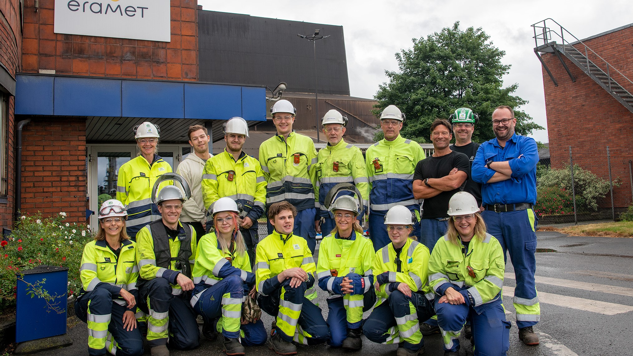 Group of people. Large group of summer workers organized as a school photo in two lines, all dressed in yellow and blue work clothes and white helmets. Outside the entrance gate to the factory premises.