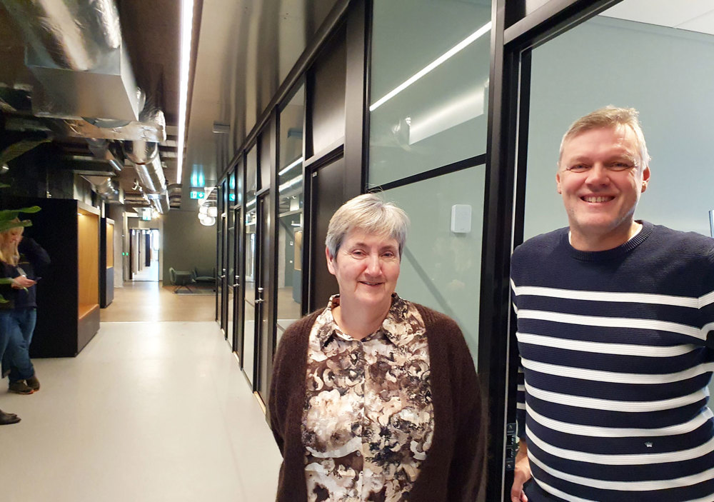 woman and man, posing, standing in an open door in a long office enviroment corridor.