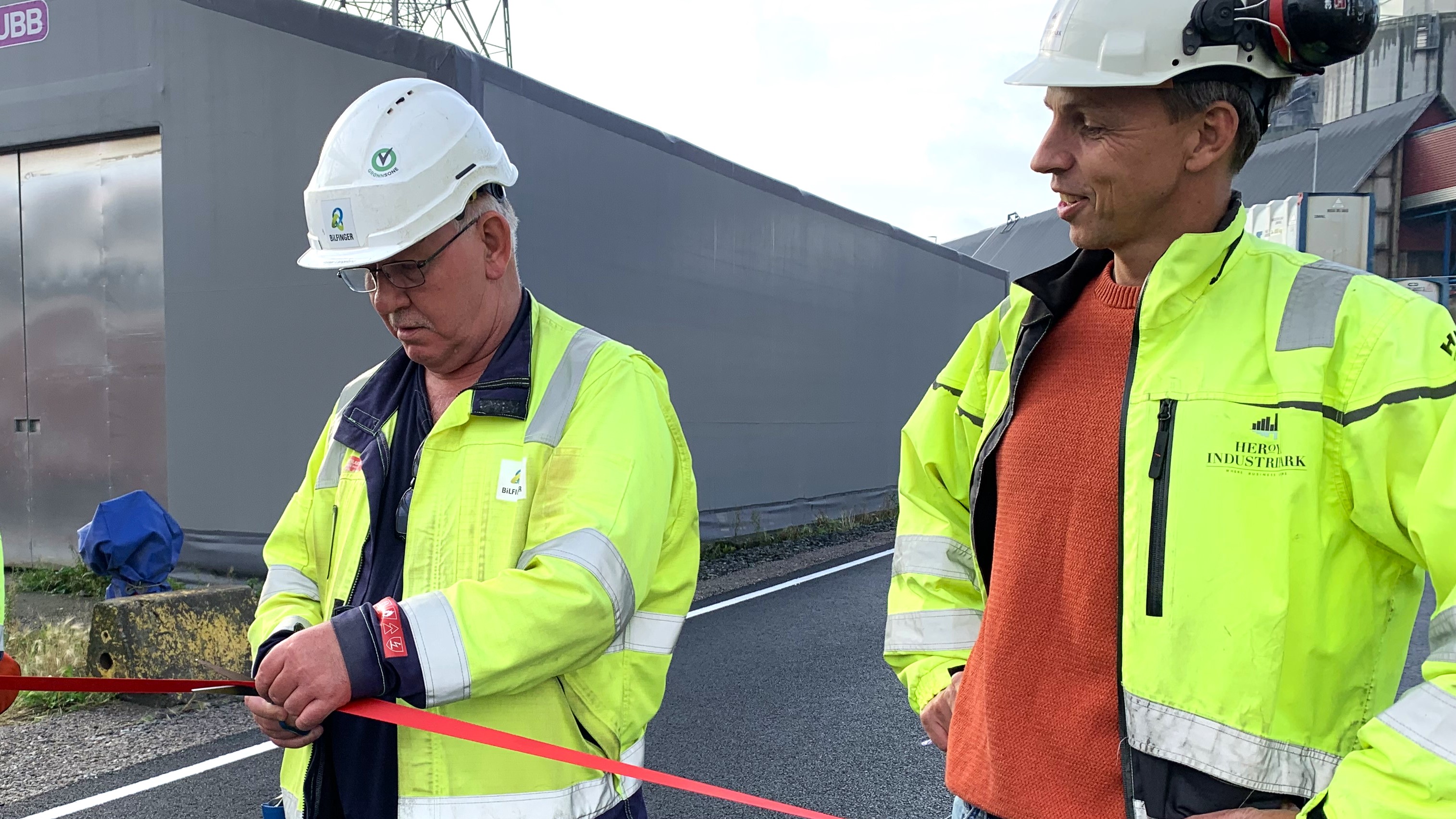 to menn i gule jakker og hvite hjelmer, den ene mannen holder en saks og er i ferd med å klippe over et rødt langt bånd, offisiell åpning. Den andre mannen har hendene i siden og ser på snorklippingen.