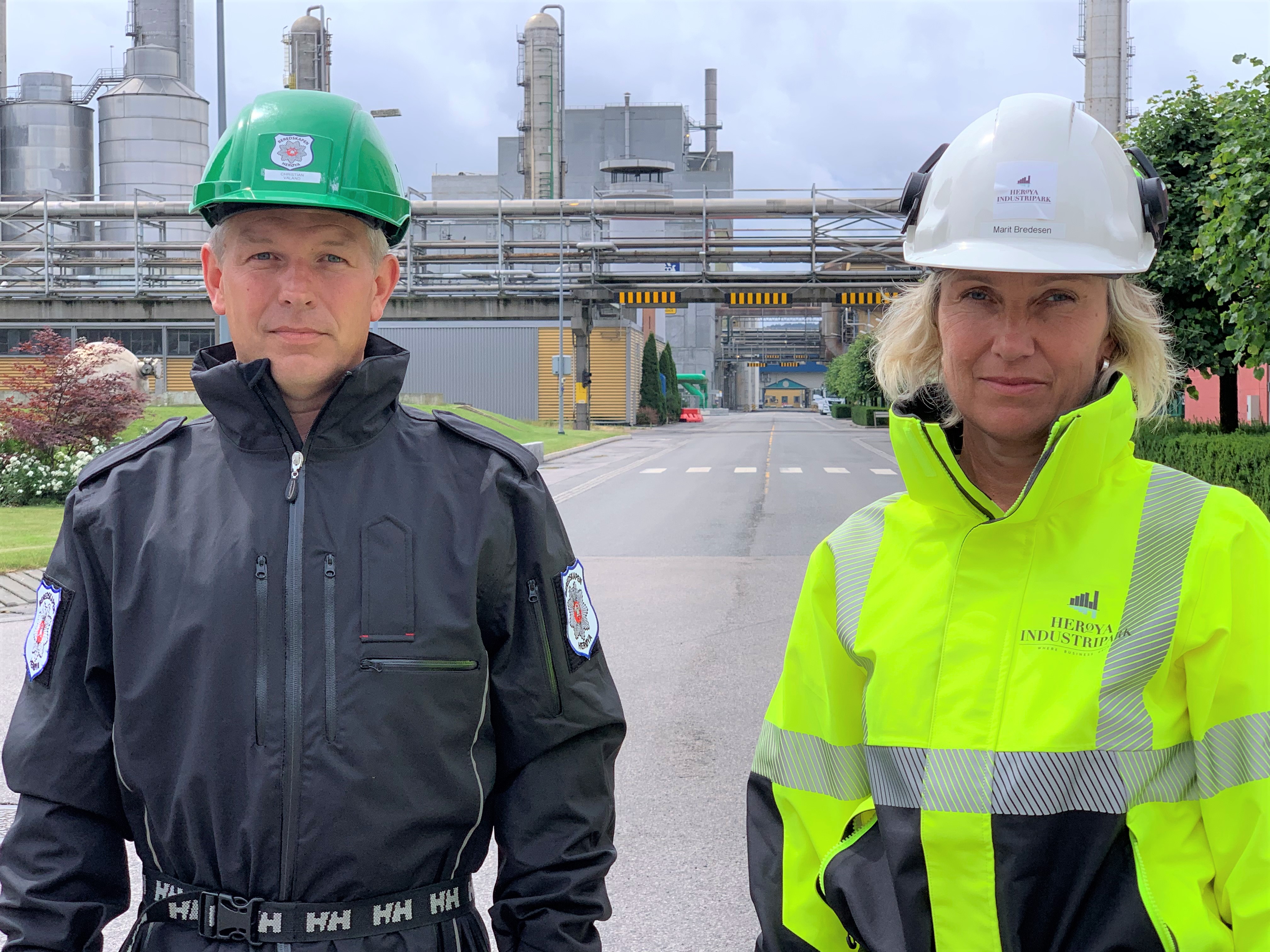 to personer, mann og kvinne står i industripark, portrett, poserer. Han kledd i mørk uniformsjakke med grønn hjelm, hun kledd i gul og blå arbeidsjakke med hvit hjelm.