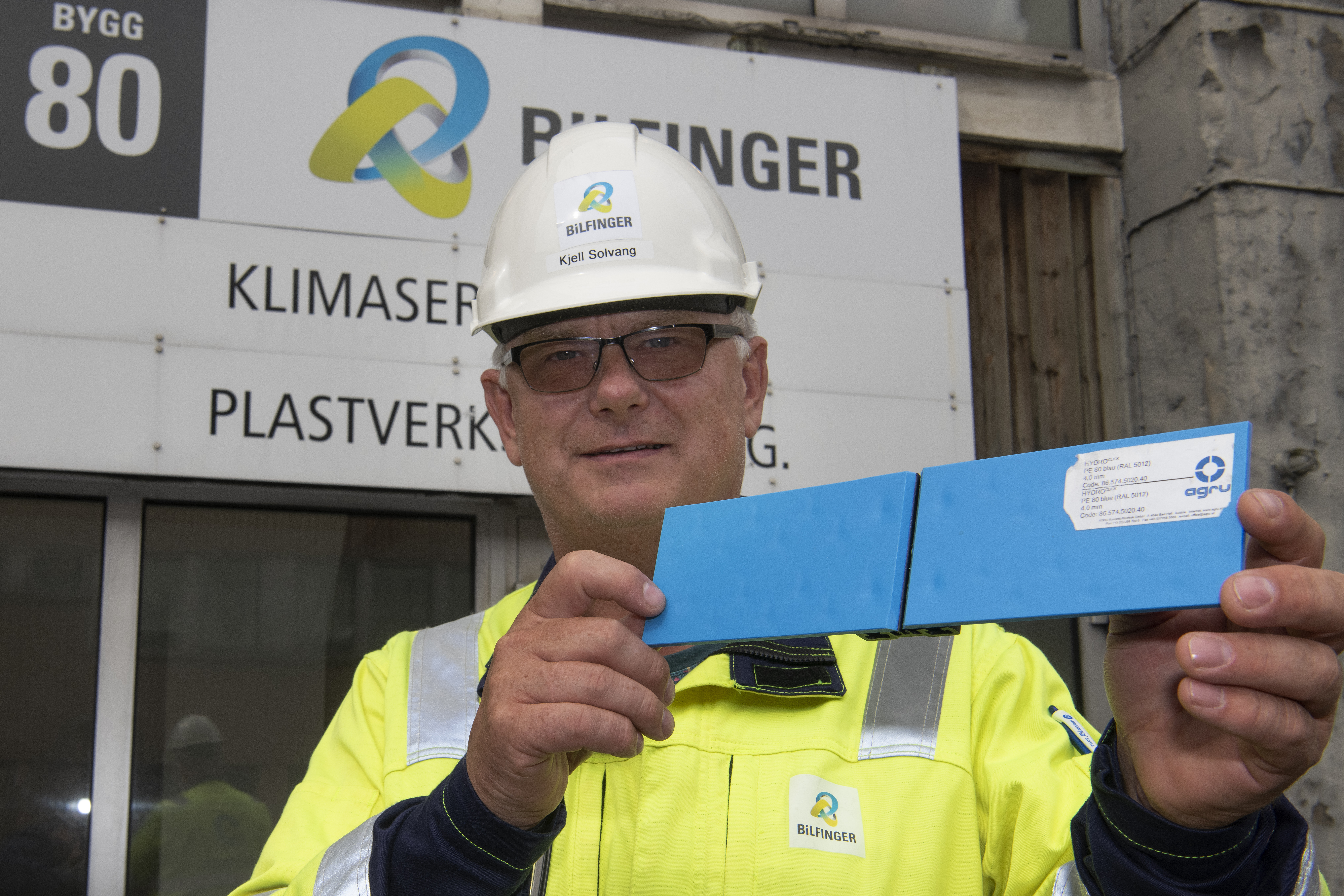 portrett, mann i gul jakke og hvit hjelm, poserer, holder opp et lite element blått plastmateriale, 