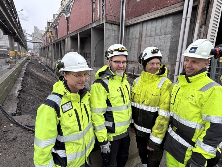 four men, posing, standing in front of a brick building, deep ditch in background.