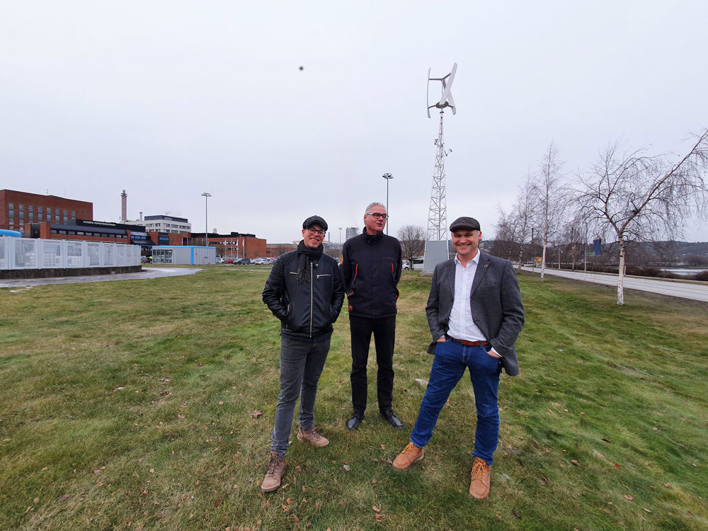 three men standing on a lawn, windmill with vertical blades in background.
