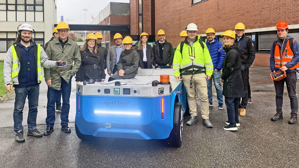 Approximately 15 people standing outside, posing, some of them leaning towards  a one meter tall autonomous blue vehicle. Industrial park, they are all wearing yellow and white helmets.