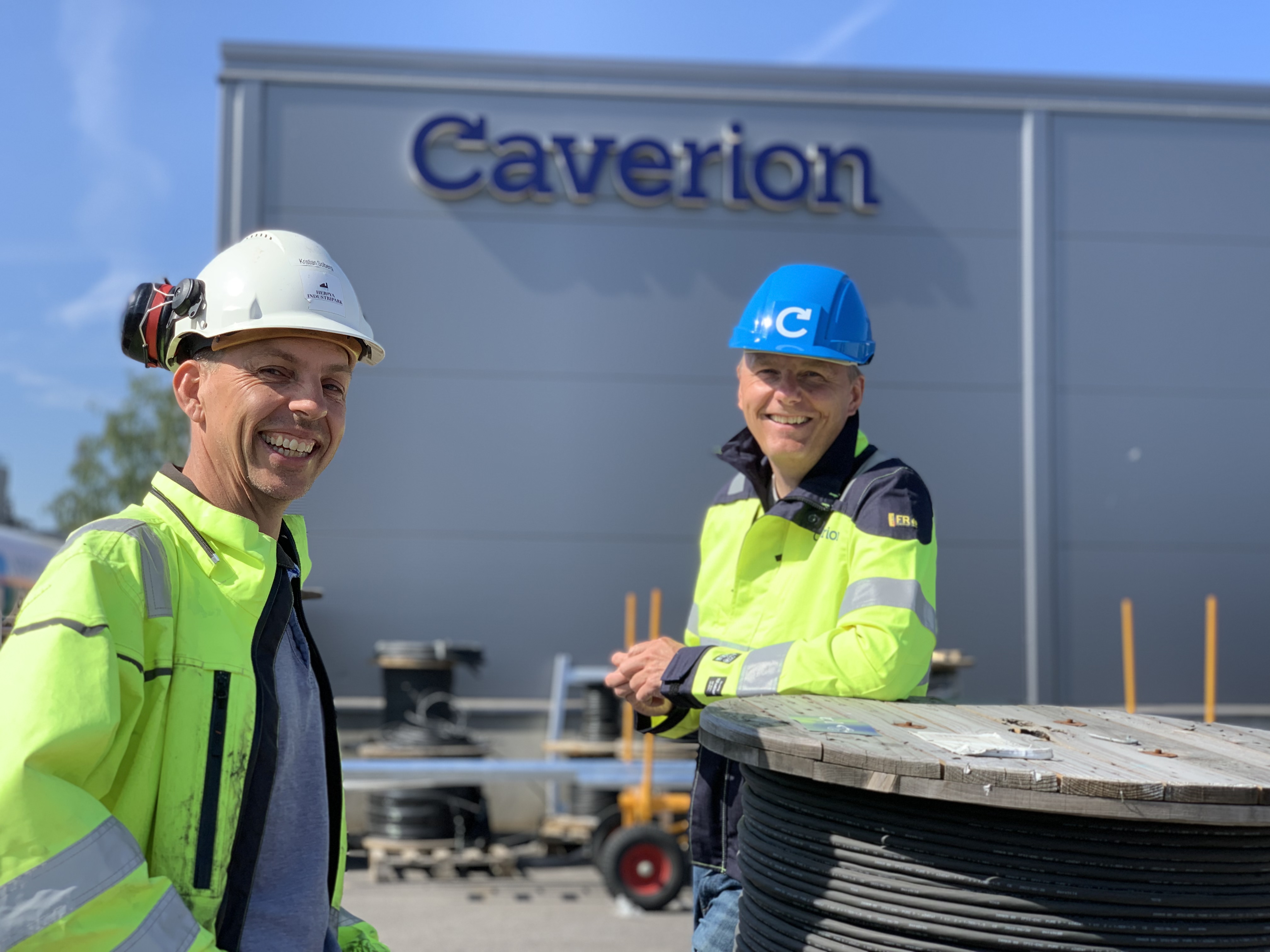 two men outside a building with a big logo on the wall. Both men wearing yellow jackets, one with blue helmet and one with white helmet.