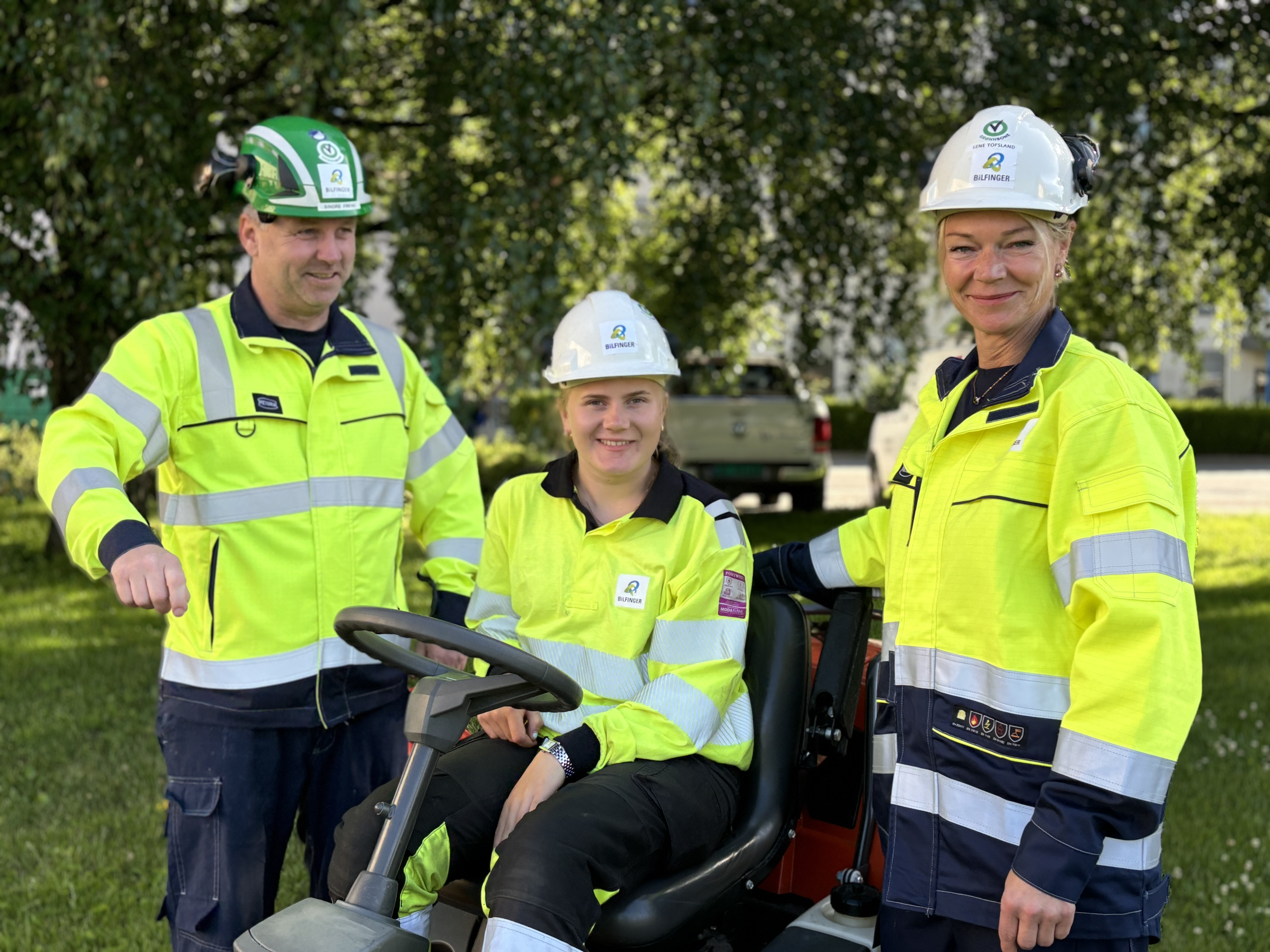 tre personer, poserer, grønt miljø, gule arbeidsjakker og hjelm, en person sitter på gressklipper.