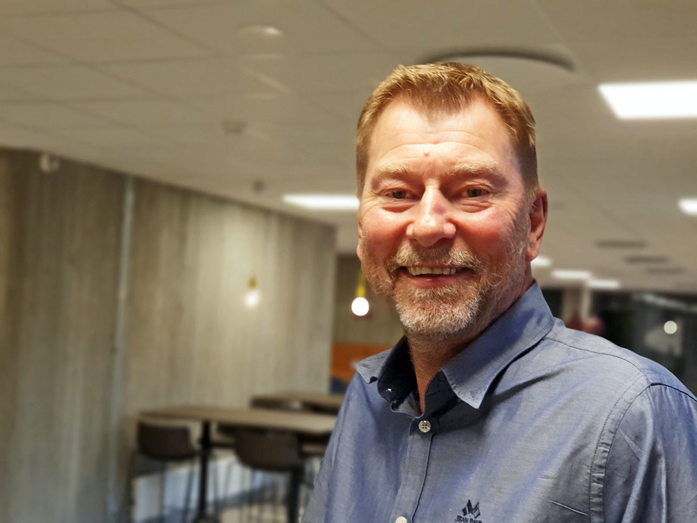 portrait of man, wearing blue shirt, standing in a big grey room.