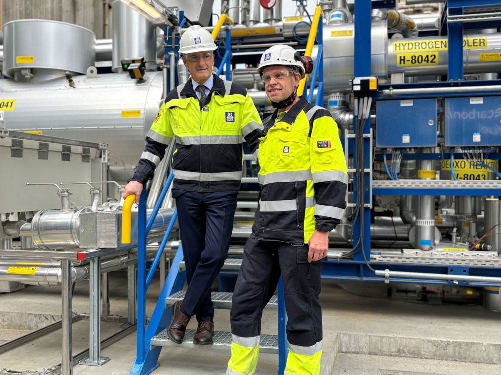 two men standing at the bottom of the stairs and in front of the hydrogen plant at Herøya. Both wearing PPE.