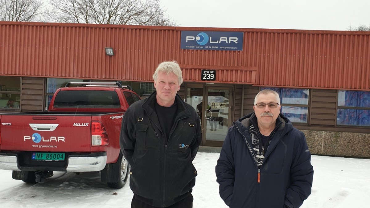 to menn står ved siden av hverandre foran et lavt verkstedbygg, med logo