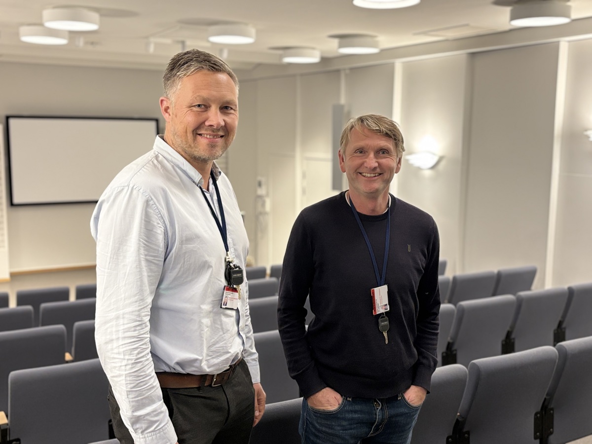 to menn poserer, auditorie, stolrader, lerret på veggen i bakgrunnen