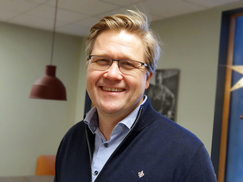 portrait of man, light blue shirt, dark blue knitted jacket, canteen furniture in background.