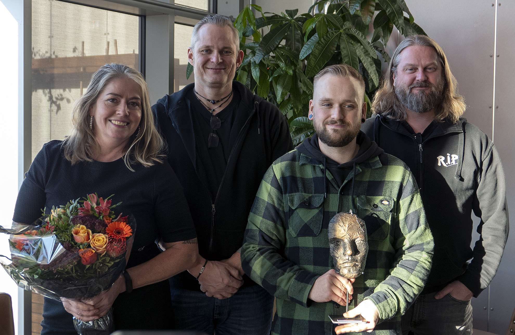 gruppe på fire personer, poserer, en holder blomsterbukett, en annen holder et lite kunstverk av et ansikt, de står i en krok med grønne høye planter. Prisvinnere.