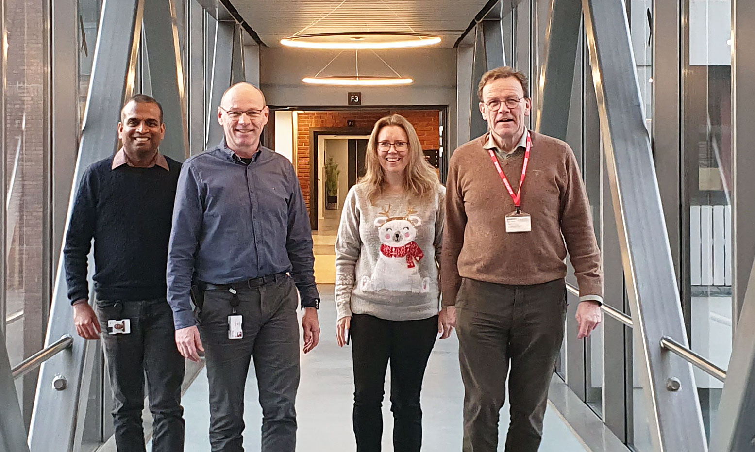 four people walking towards photographer, in a glass covered corridor/tunell, day light, research park.