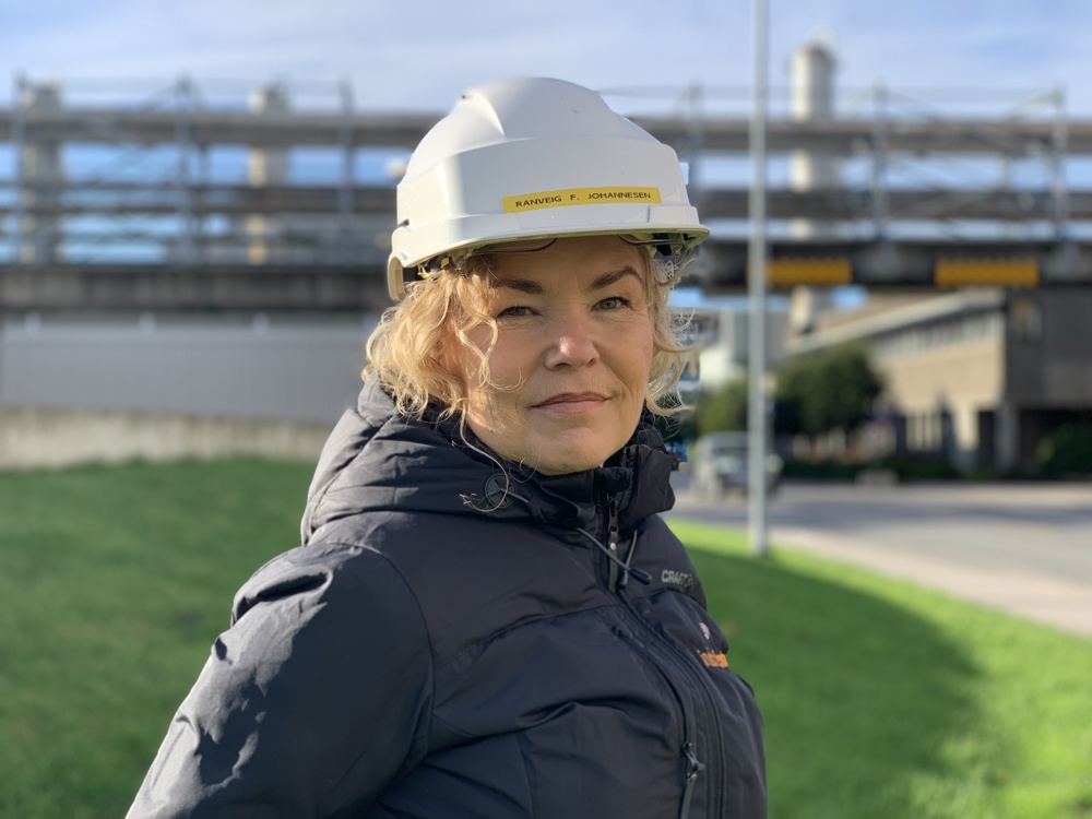 portrait, woman, blue jacket, white helmet, green lawn and industrial pipes in background.