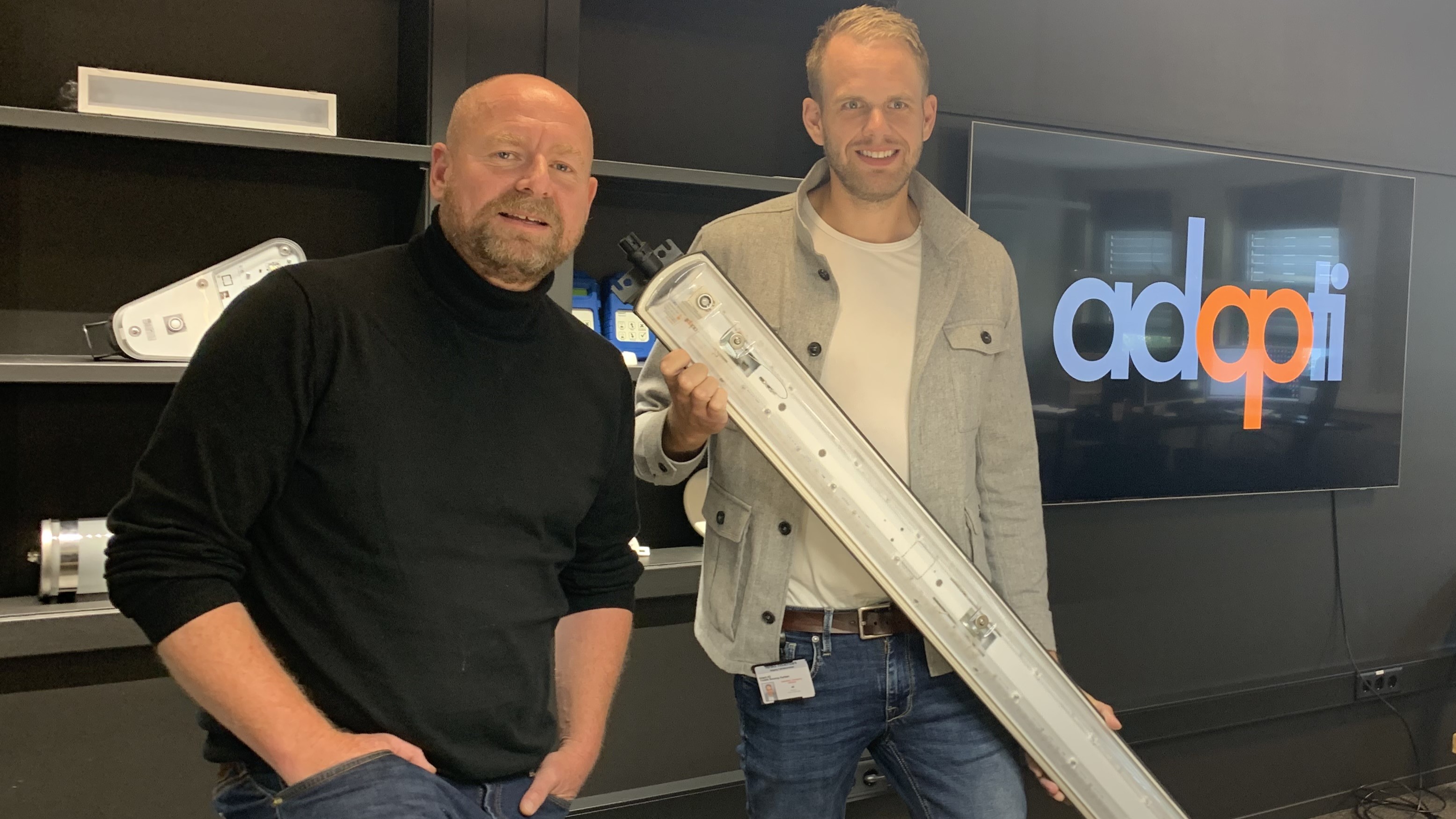 two men posing, standing in an office environment, black painted walls in background, also logo on a screen. One man holding a narrow long lamp for industry.