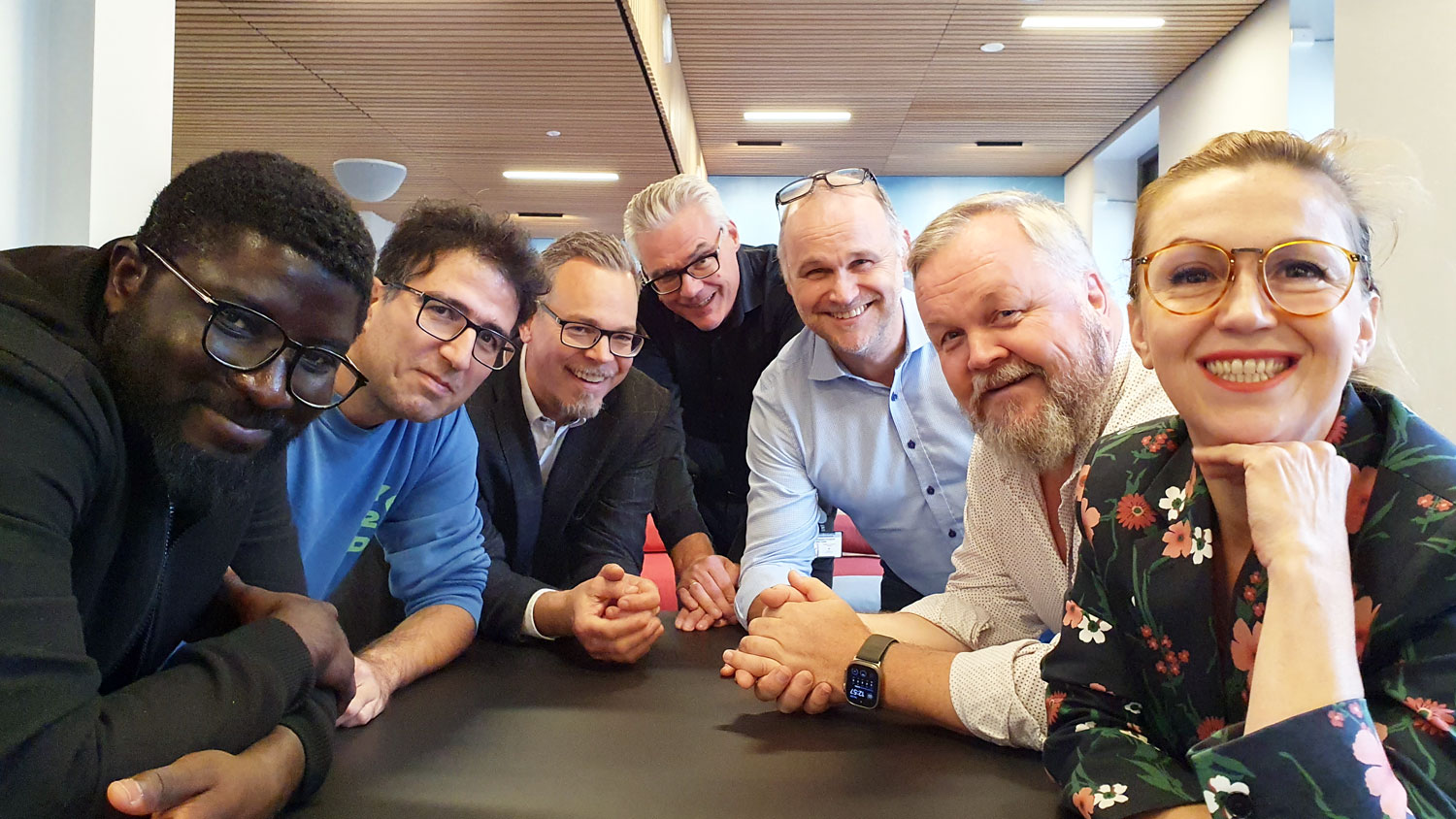 Seven people leaning into the table, standing close together, posing, office environment