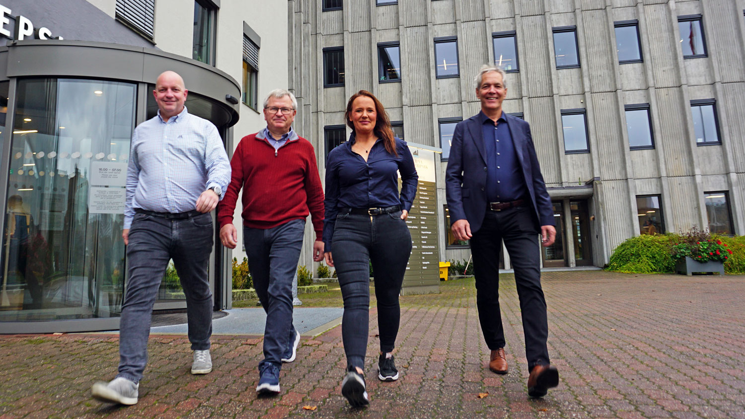 four persons walking towards the camera, posing, office building in the background