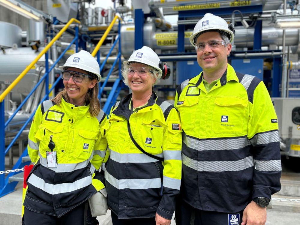 two women and one man standing in front of an electolyser, posing, smiling. all wearing PPE.